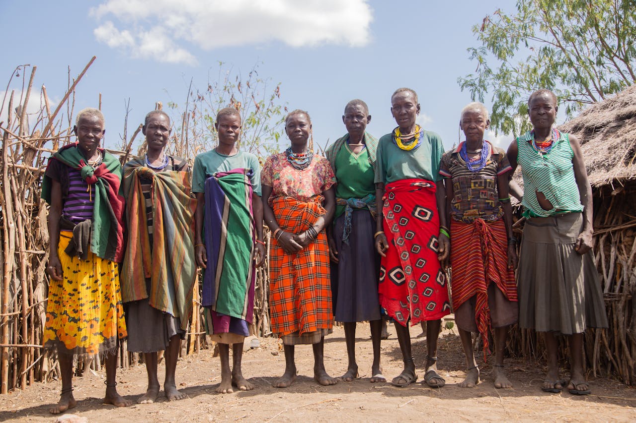 A group of women from an African tribe in traditional attire posing outdoors in their village.
